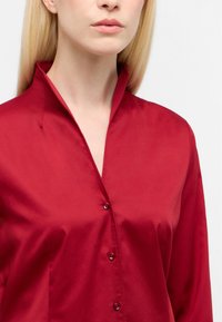 Blusa di raso rosso con colletto alto, caratterizzata da una chiusura frontale con bottoni e una texture liscia. Il design include dettagli di cucitura sottili.