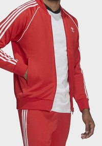 Red athletic tracksuit with white three-stripe accents on sleeves and pants. Features zip closure and two side pockets. Smooth, lightweight fabric.
