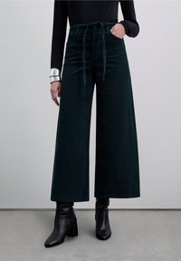 Pantalon large en velours côtelé vert foncé, avec un côtelé vertical, une taille haute ornée d'une ceinture nouée, et des poches latérales. Associé à des bottines.