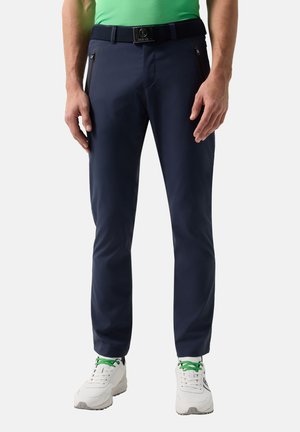 Mand iført marineblå slim-fit bukser, sort bælte, hvide sneakers med grønne snørrebånd og en grøn skjorte, stående mod en lys baggrund.