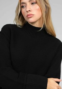 Femme blonde avec un maquillage léger portant un pull en tricot noir texturé, regardant sur le côté avec les bras croisés devant.