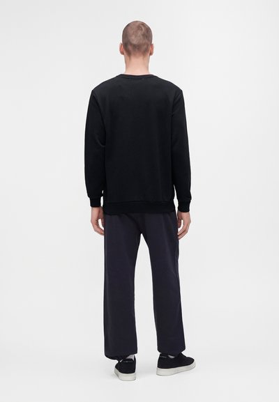 Homme debout de dos, portant une chemise noire à manches longues, un pantalon sombre et des baskets noires sur un fond blanc uni.