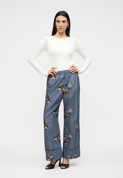 Femme aux longs cheveux foncés portant un haut blanc à manches longues, un pantalon large bleu à rayures florales, et des talons ouverts noirs, debout les mains sur les hanches.
