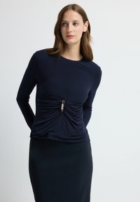 Kobieta o jasnobrązowych włosach, ubrana w granatową bluzkę z długim rękawem z marszczoną przednią częścią i dekoracyjną broszką, w połączeniu z ciemną spódnicą, na białym tle.