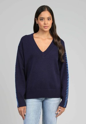 Jeune femme aux longs cheveux bruns portant un pull bleu marine en V avec des détails de couture bleus sur les manches et un jean bleu clair.