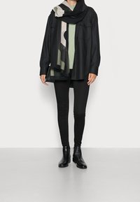 Schwarze Hemdbluse, grüner Strickpullover, schwarze Leggings, schwarze Ankle Boots. Der Schal zeigt geometrische Muster in Schwarz, Beige und Olivgrün.