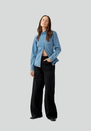 Vrouw met lang bruin haar, die een gedeeltelijk dichtgeknoopte blauwe spijkershirt en loszittende zwarte broek draagt, tegen een effen grijze achtergrond.