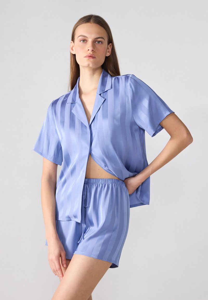 Lys blå stripete pysjamas sett laget av glatt stoff, med en kortermet skjorte som kan knytes og shorts med elastisk liv og snor.
