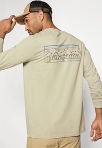 Μπεζ πουκάμισο με μακριά μανίκια που διαθέτει γραφικό σχέδιο με βουνά και τη λέξη "patagonia" σε μαύρο, με πολύχρωμες οριζόντιες ρίγες από πάνω.