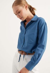 Camicia azzurra a maniche lunghe con colletto, taschino frontale e dettagli con cordoncino in vita. Abbinata a pantaloni bianchi, caratterizzati da una texture liscia e bottoni.