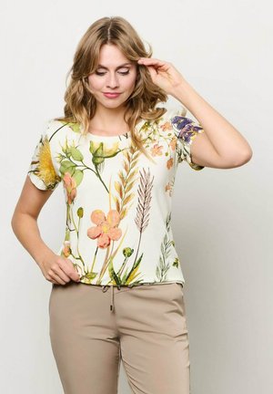 Vrouw met golvend blond haar, gekleed in beige broek en een wit shirt met korte mouwen met kleurrijke bloemen- en plantenprints, die haar haar rechtzet.