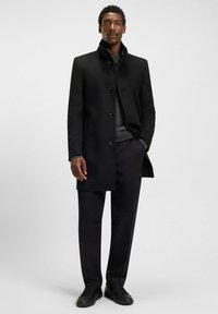 Uomo che indossa un cappotto nero con bottoni sopra un maglione grigio scuro, pantaloni neri e sneakers nere, in piedi contro uno sfondo chiaro e semplice.