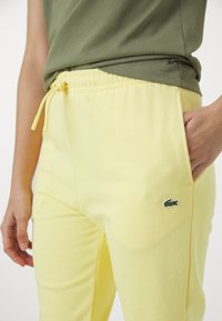 Gule joggebukser med snor i midjen, sidelommer og en liten grønn logo på venstre lår. Laget av myk, pustende stoff.
