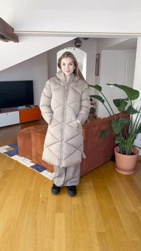 Casaco comprido acolchoado bege claro com padrão em espinha de peixe, fecho de correr e bolsos, combinado com calças cinzentas e sapatos pretos, contra um interior luminoso.