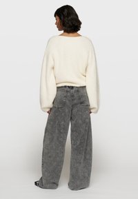 Überdimensionierter cremefarbener Pullover mit weicher Textur, kombiniert mit lässigen grauen Jeans mit einem subtilen Blumenmuster und Kontrastnähten.