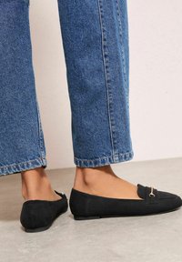 Černé semišové mokasíny s plochou podrážkou, zdobené malým zlatým akcentem vpředu, kombinované s modrými širokými džíny.