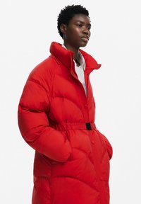 Abrigo acolchado rojo con cuello alto, cintura ceñida y cierre de botones. Presenta un diseño de acolchado cosido y bolsillos laterales. Textura suave.