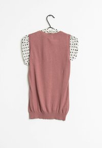 Mouwloos kledingstuk met een roze lichaam en korte witte stippenmouwen. Zachte textuur met ribbelboord aan de taille. Hangt aan een zwarte haak.
