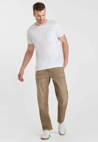 Homme marchant, portant un t-shirt blanc à manches courtes avec une poche poitrine, un pantalon marron clair et des baskets blanches sur un fond clair et uni.