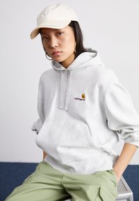 Light gray hooded sweatshirt with front pocket, featuring a small embroidered logo. Paired with light green pants and a beige cap.