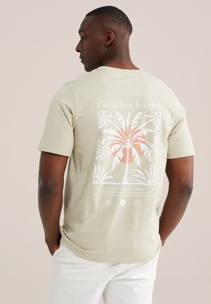 Mężczyzna noszący beżowy T-shirt z tropikalną palmą i napisem "Paradise Found" na plecach, w połączeniu z białymi spodniami.