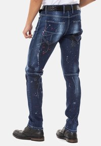 Blauwe spijkerbroek met een slim fit, voorzien van spetterpatronen in rood, groen en wit; vijf zakken en een zwarte riem.