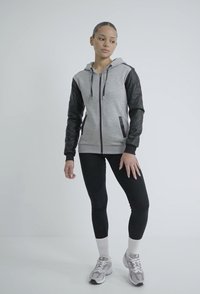 Sivi zip-up dukserica s crnim kvadratnim rukavima, prednje džepove, nošena s crnim tajicama i sivim patikama. Lagani materijal i ležeran dizajn.