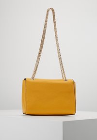 Žlutá syntetická kabelka s pevnou strukturou. Má zlatý řetízkový popruh a texturovaný povrch s černými akcenty podél okrajů.