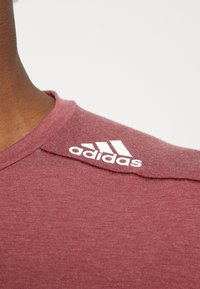 Bordová tričko Adidas s kulatým výstřihem, vyrobené z texturované tkaniny a s bílým logem na rameni. Hladký povrch, krátké rukávy.