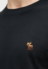 Κοντινή λήψη μαύρου πουκάμισου με μικρό κεντημένο καφέ ελάφι με κίτρινα κέρατα στο στήθος κοντά στον λαιμό.