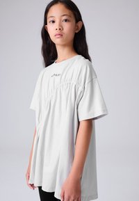 Svijetlosiva široka majica kratkih rukava, s naboranima sa strane i izvezenim tekstom na prsima. Glatka tekstura tkanine.