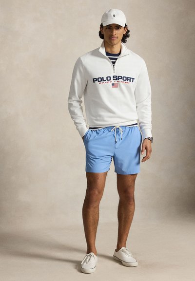 Νέος άνδρας φοράει λευκό φούτερ Polo Sport, ανοιχτό μπλε σορτς, λευκό καπέλο και λευκά αθλητικά παπούτσια, στέκεται μπροστά σε ουδέτερο φόντο.