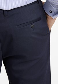 Marineblauwe op maat gemaakte broek van een zachte stof, voorzien van een achterzak met knoopsluiting en subtiele stikseldetails.