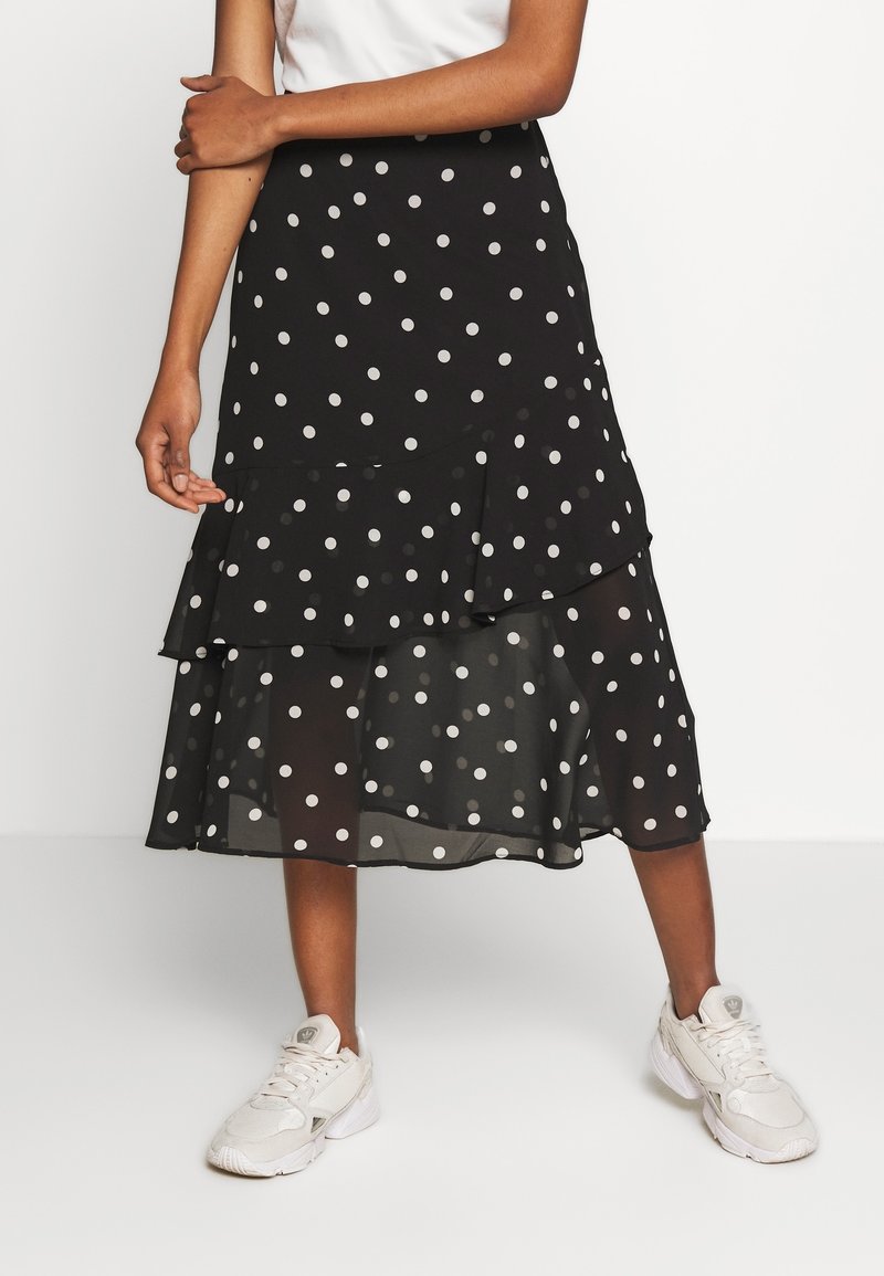 Femme portant une jupe midi noire à pois blancs et des baskets blanches, debout avec un bras croisé et un bras détendu.
