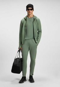 Athletic outfit in olive green, consisting of a fitted top, zip-up hoodie, and tapered pants. Includes black accessories and a sports bag.