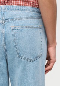 Κοντινό πλάνο ενός ατόμου που φοράει ανοιχτό μπλε τζιν παντελόνι και κόκκινο καρό πουκάμισο, εμφανίζοντας την πίσω τσέπη και μέρος του δεξιού τους χεριού.