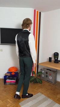 Vihreät räätälöidyt housut ja musta satiininen liivi valkoisen paidan päällä. Tummat kengät ja valkoiset sukat. Moderni sisustus, jossa värikkäitä seinäraitoja.