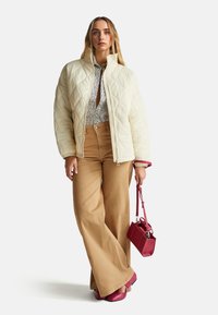 Prošívaná krémová bunda s vysokým límcem, vzorovaná košile, široké kalhoty v béžové barvě a burgundská kabelka. Obuv je v odpovídající burgundské barvě.