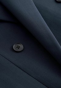 Tissu de costume bleu marine foncé avec un motif chevrons fin, doté d'un seul bouton noir à quatre trous et d'un revers plié.
