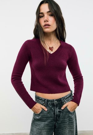 Donna che indossa un maglione corto a V viola, collane d'oro a strati con ciondoli a forma di cuore e fiore, e jeans neri a vita alta con le mani nelle tasche.