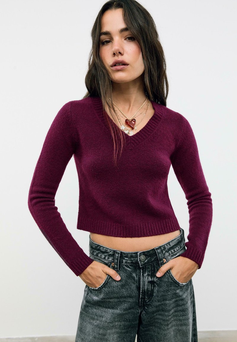 Femme portant un pull violet coupé à décolleté en V, des colliers en or superposés avec des charms en forme de cœur et de fleur, et un jean noir taille haute délavé avec les mains dans les poches.