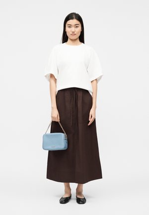 Nuori nainen valkoisessa lyhythihavaisessa paidassa, pitkäsäihkässä tummanruskeassa hameessa, mustissa ballerinoissa, pitäen kädessään pientä vaaleansinistä ketjullista käsilaukkua.