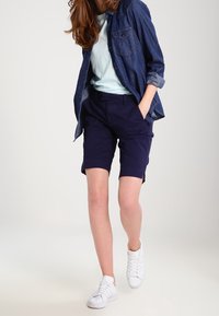 Shorts bleu foncé avec une coupe ajustée, associés à un t-shirt bleu clair et une chemise en denim à boutons. Des baskets blanches complètent la tenue.
