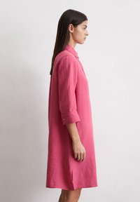 Rozā lina krekls-vakarkleita ar apkakli, sarullētām piedurknēm un sānu šķēlum. Tam ir pogas gar priekšpusi, lai aizvērtu, un brīva piegriezuma dizains.