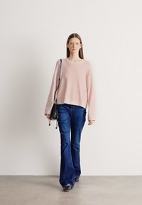 Ροζ ανοιχτόχρωμο πουλόβερ με χαλαρή πλέξη και στρογγυλή λαιμόκοψη, φορεμένο πάνω από σκούρα μπλε τζιν καμπάνες. Μια μικρή μαύρη τσάντα κρέμεται από τον ώμο.