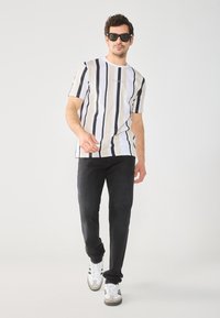 Short-sleeve striped t-shirt in white, black, and beige. Paired with black pants and white sneakers. Model wears dark sunglasses.