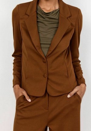 Femme portant un blazer marron sur mesure et un pantalon assorti, les mains dans les poches, par-dessus un haut drapé vert olive.