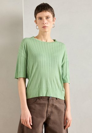 Világos zöld kötött pulóver rövid ujjakkal, függőleges bordázott textúrával és laza szabással. Barna, lazán illeszkedő nadrággal párosítva.