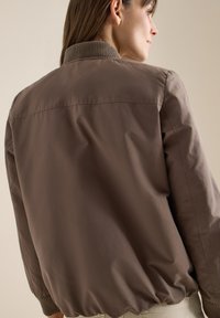 Femme portant une veste bomber marron avec col et poignets côtelés, vue de dos sur un fond neutre.