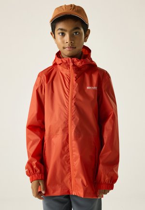 Niño con chaqueta de lluvia naranja con capucha y gorra marrón, mirando hacia adelante sobre un fondo claro y liso.